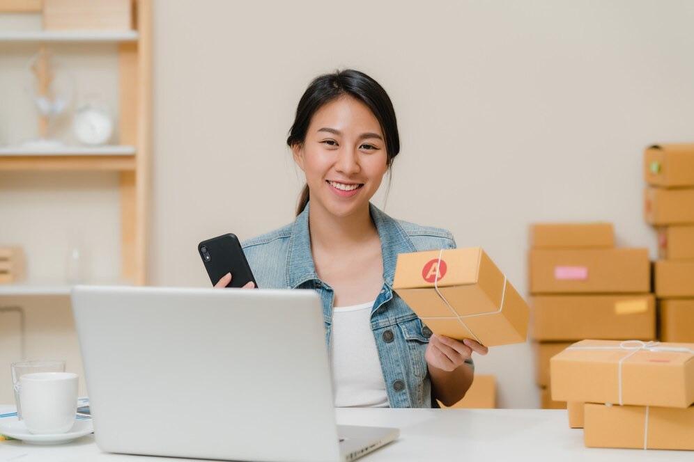 Smiling online store owner holding a parcel and her phone at her laptop